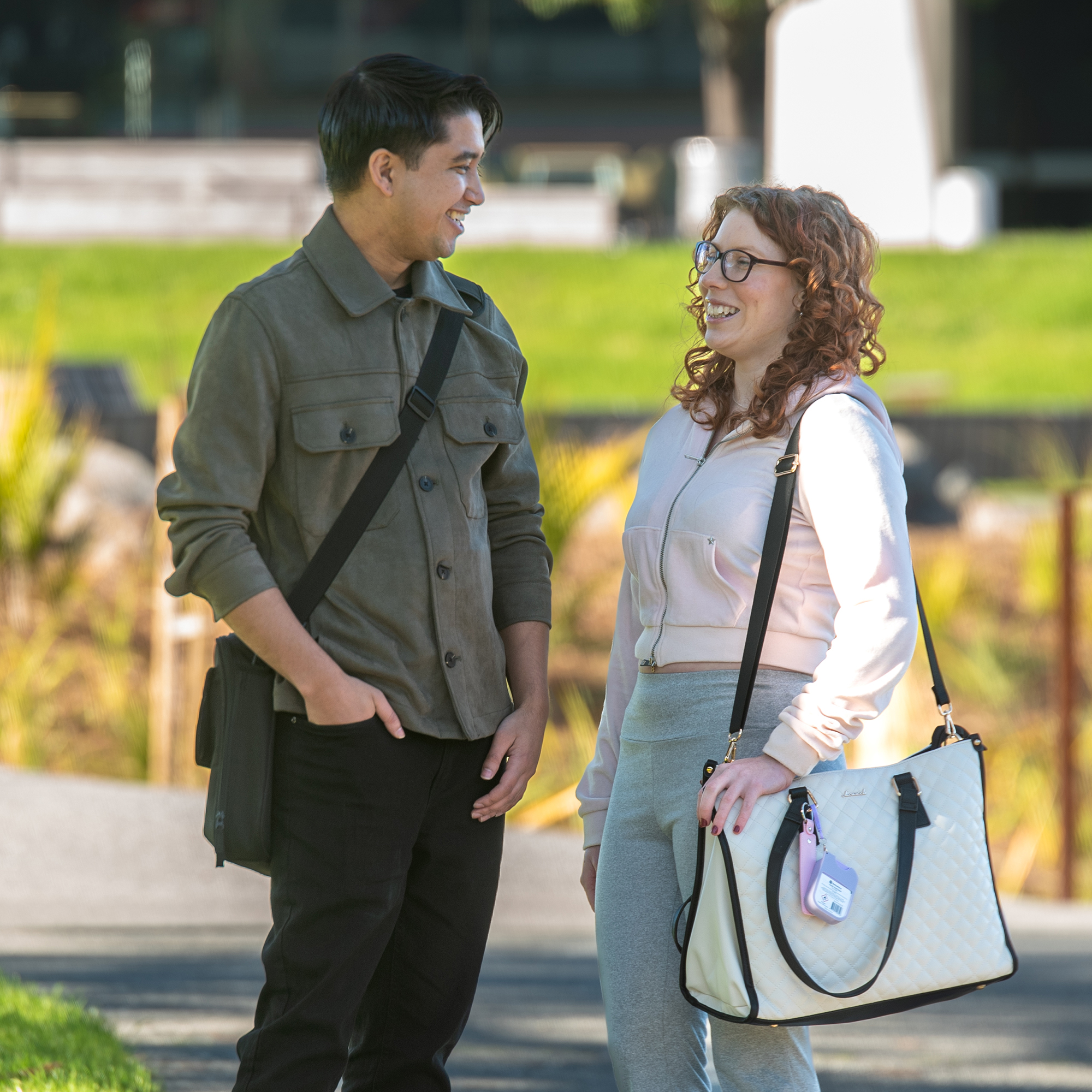 Two people stand on a paved path in a park, facing each other in conversation. One wears a green jacket, black pants, and a black shoulder bag. The other wears a light pink hoodie, gray pants, and carries a large cream quilted bag. Green grass and plants fill the background